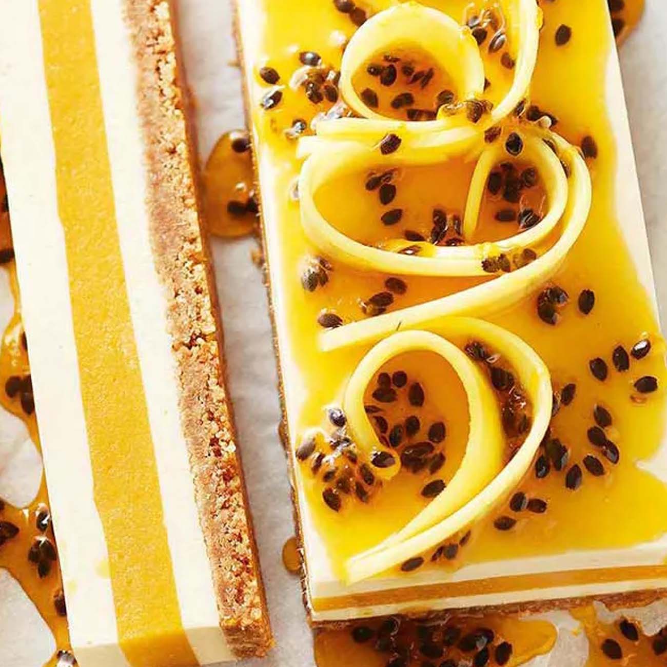 A layered mango and passionfruit slice with a biscuit base, topped with fresh passionfruit pulp and curls of fresh mango.