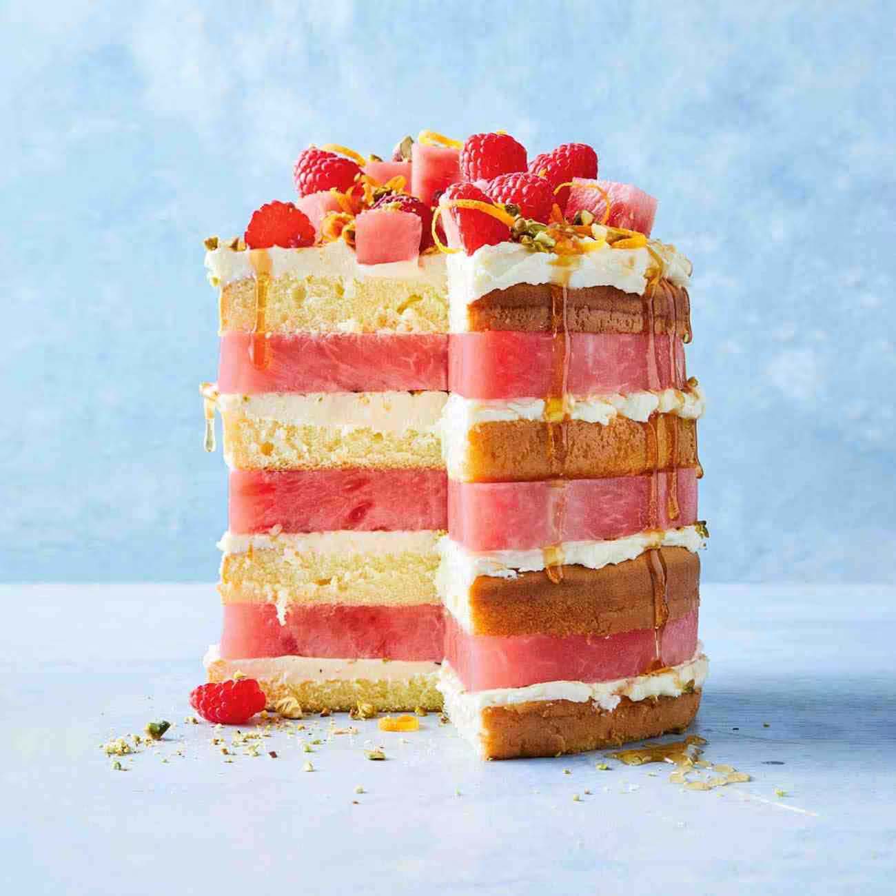 A tall, tiered layer cake stacked with thick slices of fresh watermelon and cream, topped with raspberries and a syrup drizzle.