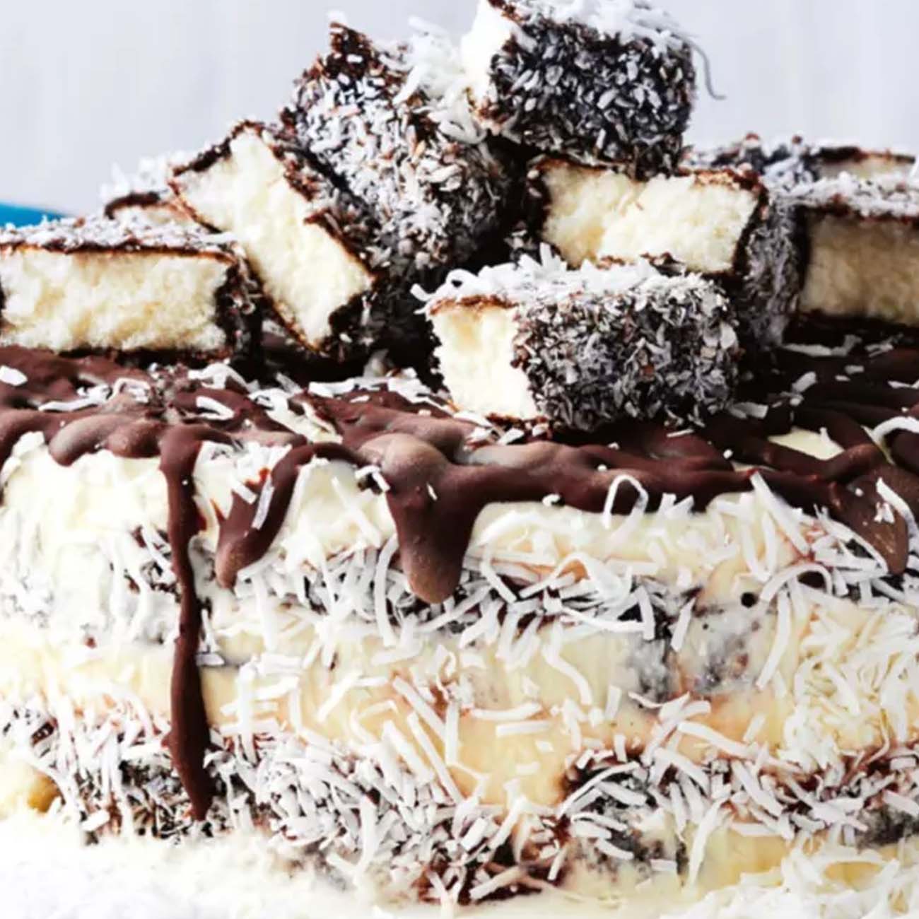 A lamington celebration cake covered in whipped cream and shredded coconut, topped with cubed mini lamingtons and a chocolate drizzle.