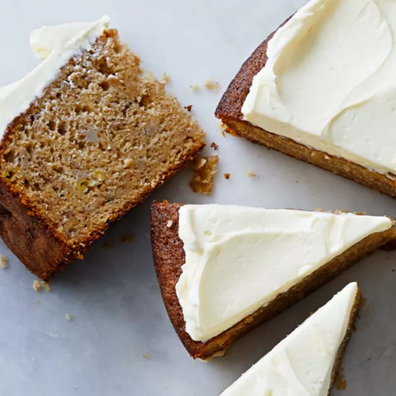 A round banana or carrot cake with thick white frosting, shown cut into slices on a light-coloured marble board.