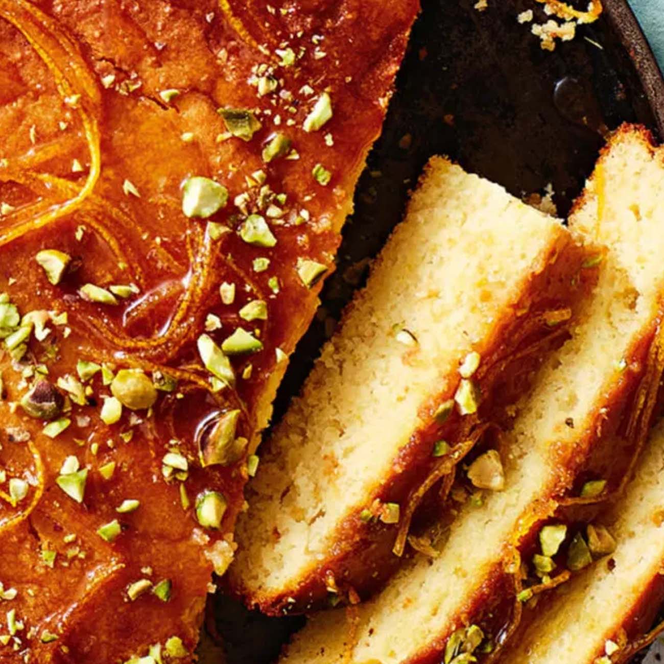 A moist orange cake drenched in syrup, topped with crushed pistachios, with a few slices cut to reveal the inside texture.
