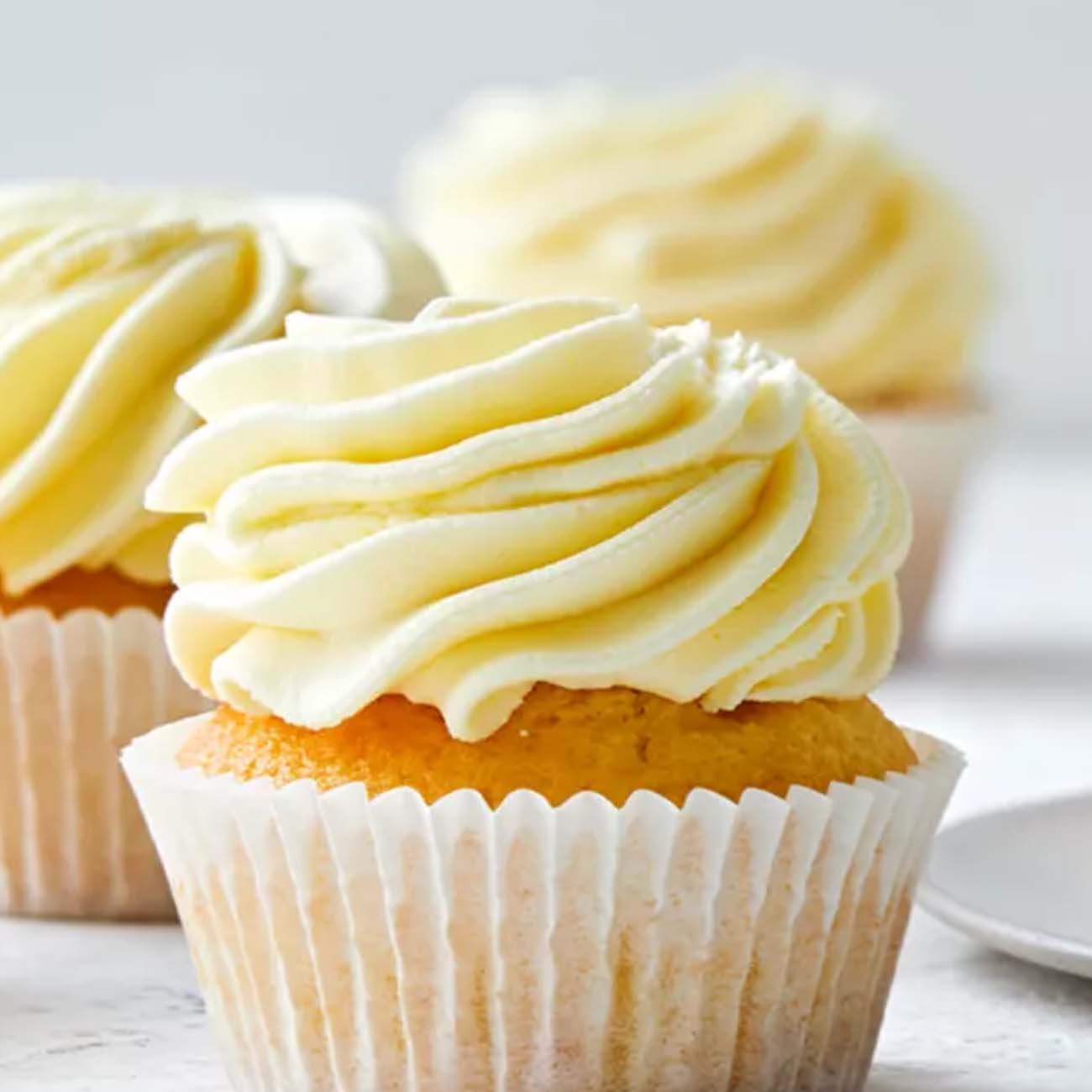Close-up of golden vanilla cupcakes topped with thick, high swirls of smooth buttercream icing in white paper cases.